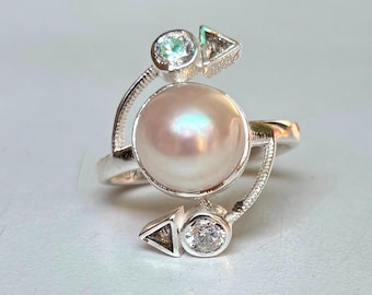 May include: A silver ring with a large, iridescent pearl at its centre. The ring has an unusual, asymmetrical design with small, clear accent stones and geometric shapes. The pearl has a soft, pinkish colour.