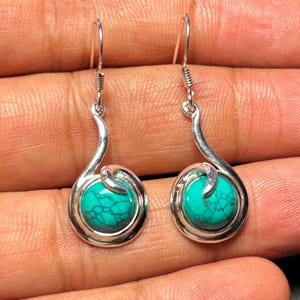 May include: A pair of silver-toned earrings with turquoise-colored, round stones. The earrings have a curved design with a spiral detail above the stone. The turquoise stones have a mottled appearance.