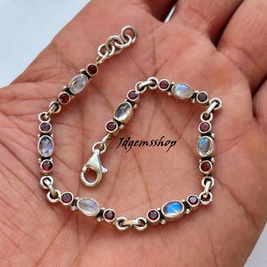 May include: A silver chain bracelet featuring alternating oval moonstones and small round red garnet gemstones. The bracelet includes a lobster claw clasp and a short extender chain with several links. The text Jdgemsshop is visible.