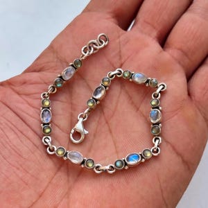 May include: A silver bracelet with a series of oval and round gemstones. The bracelet alternates between clear, blue, and green-toned stones. The clasp is a small silver lobster claw. The bracelet is displayed on a light-coloured surface.