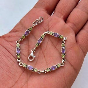 Op de afbeelding: Zilveren armband met afwisselend ovale paarse en ronde groene edelstenen. De armband heeft een karabijnsluiting en is ongeveer 17,78 cm lang.