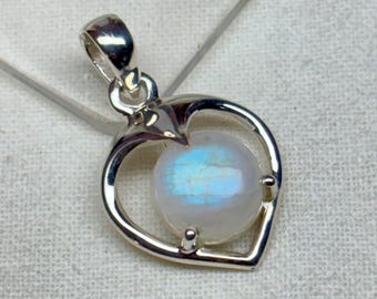 May include: A silver pendant with a heart-shaped frame. The pendant holds a round, iridescent moonstone. The moonstone has a blue and white glow. The pendant is on a white surface.