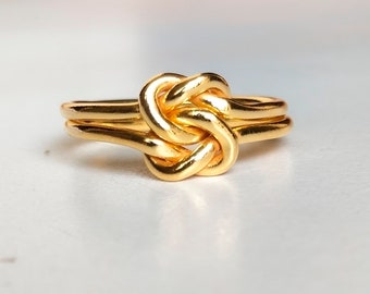 Puede incluir: Un anillo dorado con un diseño de nudo. El anillo presenta una doble banda y un nudo prominente en el centro, creando un aspecto único y elegante. El anillo está hecho de un material metálico brillante.