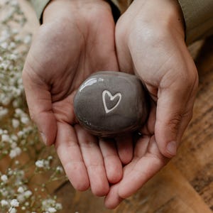 Puede incluir: Una piedra gris pulida y lisa con un contorno de corazón blanco grabado en el centro. La piedra tiene un acabado brillante y una forma orgánica redondeada, descansando en las palmas de las manos, mostrando su tamaño compacto.