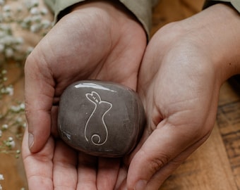 Puede incluir: Una piedra gris pulida y lisa que presenta un dibujo de líneas blancas con la silueta de un conejo. La piedra es redondeada y compacta, con el diseño del conejo centrado en su superficie, mostrando líneas limpias sobre el fondo gris.