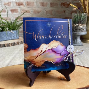 Könnte beinhalten: Eine dunkelblaue Karte mit goldener und violetter abstrakter Kunst und dem Text "Wunscherschiller" auf einem Holzständer.