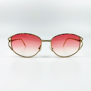 Puede incluir: Gafas de sol ovaladas doradas con lentes de degradado rojo. Las gafas tienen un diseño delicado e intrincado en el marco.