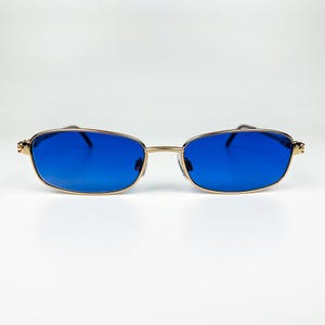 May include: Gold-framed sunglasses with rectangular blue lenses. The glasses have a thin gold frame and a small bridge. The temples are also gold. The sunglasses are set against a white background.