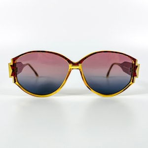 Puede incluir: Un par de gafas de sol vintage con montura roja y dorada y lentes degradados. Las lentes son de color azul oscuro en la parte inferior y se desvanecen a un color rosa claro en la parte superior.