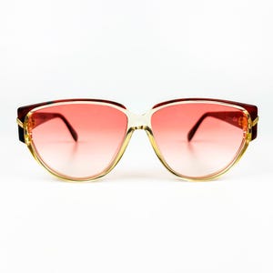 Puede incluir: Gafas de sol vintage de gran tamaño con lentes rosas degradadas. Las monturas son una combinación de transparente, dorado y burdeos oscuro. Las gafas de sol tienen una forma retro y angular, sobre un fondo blanco.