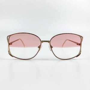 Puede incluir: Un par de gafas de color dorado con lentes de degradado rosa. Las gafas tienen una forma cuadrada con bordes redondeados.