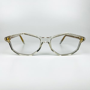 May include: Clear, geometric-shaped spectacles featuring a transparent frame with a subtle angular design. The upper rims are embellished with a row of small, sparkling clear crystals along the top edge, extending towards the temples.