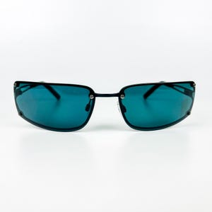 Op de afbeelding: Zonnebril met zwart montuur en teal glazen. De rechthoekige glazen zijn licht gebogen en met kleine zilveren klinknagels aan het montuur bevestigd. De zonnebril heeft een strak, modern design.