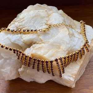Op de afbeelding: Een gouden ketting met een hangend ontwerp van donkerrode strass steentjes. De ketting ligt op een wit rotsoppervlak.