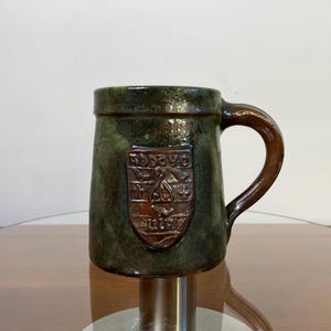Peut inclure: Une chope en céramique verte et brune avec une anse. La chope a un blason brun avec le texte "Rodbaye" et un motif d'oiseau.