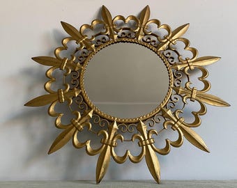 Puede incluir: Un espejo de metal dorado con un diseño de sol. El espejo está rodeado por un marco decorativo con motivos de flor de lis.