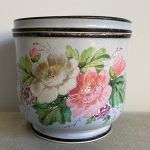 Può includere: Fioriera in ceramica azzurra con un design floreale. La fioriera presenta fiori dipinti a mano in tonalità di rosa, bianco e verde. Il bordo e la base sono accentuati da finiture nere. La fioriera è alta circa 25 cm.