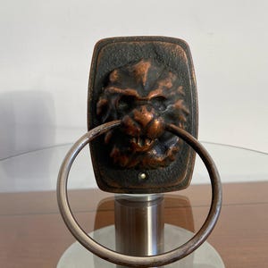 May include: A bronze-colored wall-mounted towel ring with a lion's head design. The ring is made of metal and has a smooth, polished finish.