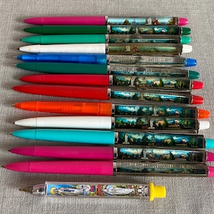 Può includere: Una collezione di penne colorate con fusti trasparenti che mostrano scene di paesaggi in miniatura. Le penne hanno cappucci e corpi di vari colori, tra cui rosa, verde, bianco, blu, rosso e arancione. Una penna è più corta con un'estremità gialla.