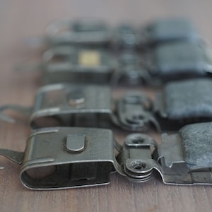 May include: A row of five silver metal clips with grey fabric pads. The clips have a hook on one end and a spring mechanism on the other. The clips are arranged in a line, with the hook end facing the viewer.
