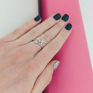 Puede incluir: Un anillo plateado con un peque&ntilde;o avi&oacute;n y una gema redonda y transparente. El anillo se lleva en un dedo, sobre un fondo rosa y blanco. El dise&ntilde;o del avi&oacute;n es detallado y la gema brilla.