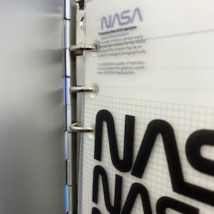 May include: A close-up of a binder with a clear plastic page showing the NASA logo and text that reads "Reproduction Art:Logotype". The text below the logo explains that the artwork can be reproduced and enlarged photographically. It also provides contact information for obtaining additional supplies of reproduction art.