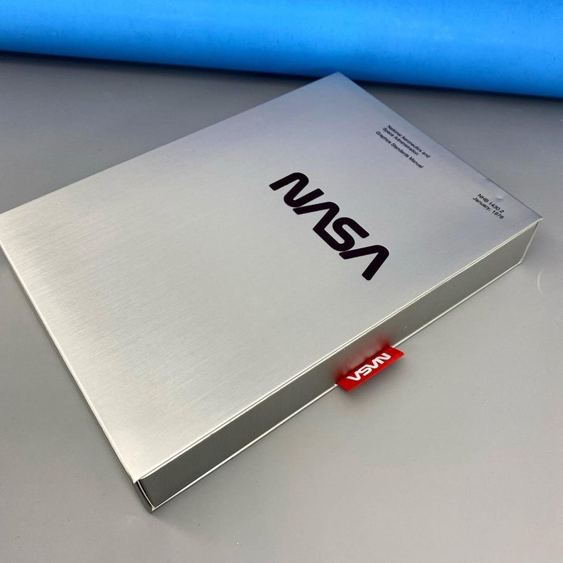 May include: A silver box with the NASA logo printed on the top. The box has a red tag with the word "NASA" printed on it.