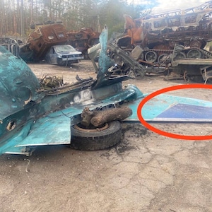 May include: A close-up of a destroyed aircraft fuselage, with a large piece of the wing lying on the ground. The aircraft is painted in a light blue color, and the wing is a darker blue. The aircraft is surrounded by debris and other destroyed vehicles.