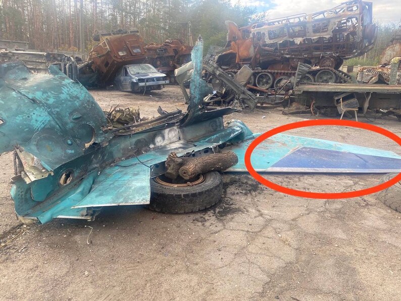 May include: A close-up of a destroyed aircraft fuselage, with a large piece of the wing lying on the ground. The aircraft is painted in a light blue color, and the wing is a darker blue. The aircraft is surrounded by debris and other destroyed vehicles.