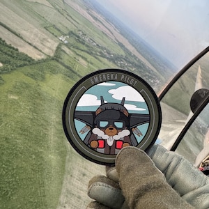 May include: A circular patch with the text "SMEREKA PILOT" around the edge. The patch features a cartoon bear wearing aviator goggles and a flight suit, with a jet plane design in the background. The patch is held in a gloved hand.