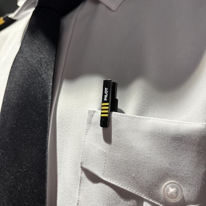 Könnte beinhalten: Ein schwarzer Pilot-Anstecker mit gelben Streifen, der an einer weißen Hemdtasche befestigt ist. Auf dem Anstecker steht in weißer Schrift "PILOT". Im Hintergrund ist eine schwarze Krawatte zu sehen.