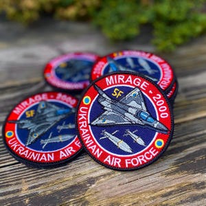 Pode incluir: Um close-up de vários patches bordados com um jato de combate com o texto "MIRAGE 2000" e "UKRAINIAN AIR FORCE".