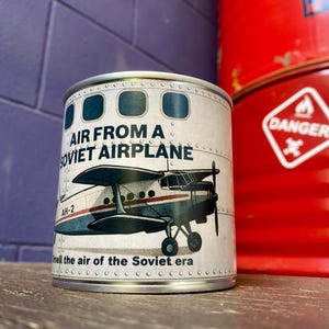 Könnte beinhalten: Eine zylindrische Konservendose mit einem Vintage-Flugzeugdesign. Die Dose ist mit "AIR FROM A SOVIET AIRPLANE" beschriftet und zeigt eine Illustration eines Doppeldeckers. Der Text "Smell the air of the Soviet era" ist ebenfalls auf der Dose aufgedruckt.