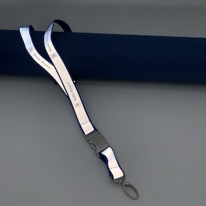 Peut inclure: Un cordon réfléchissant avec le logo et le texte Rolls-Royce. Le cordon est principalement blanc avec une bordure bleue, une attache en plastique noir et un crochet en métal argenté.