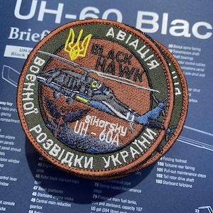 May include: A circular embroidered patch featuring a black helicopter graphic, the text "BLACK HAWK" and "SIKORSKY UH-60A", and Ukrainian Cyrillic text. The design includes a yellow trident symbol, blue and yellow accents, and a brown background.