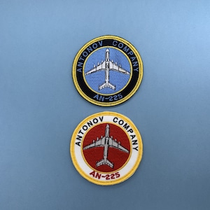 Puede incluir: Dos parches bordados con borde amarillo, fondo negro y rojo y un avión blanco. El texto en los parches dice "Antonov Company AN-225".