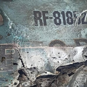 May include: Close-up of a damaged aircraft fuselage with the registration number "RF-81872" visible. The metal is heavily damaged and charred, with visible holes and cracks.