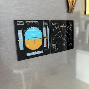 May include: Two black rectangular magnets with aviation instrument panel designs. The left magnet displays a flight instrument with blue, yellow, and white elements. The right magnet shows a curved gauge with numbers and a needle. A small elephant figurine is visible.