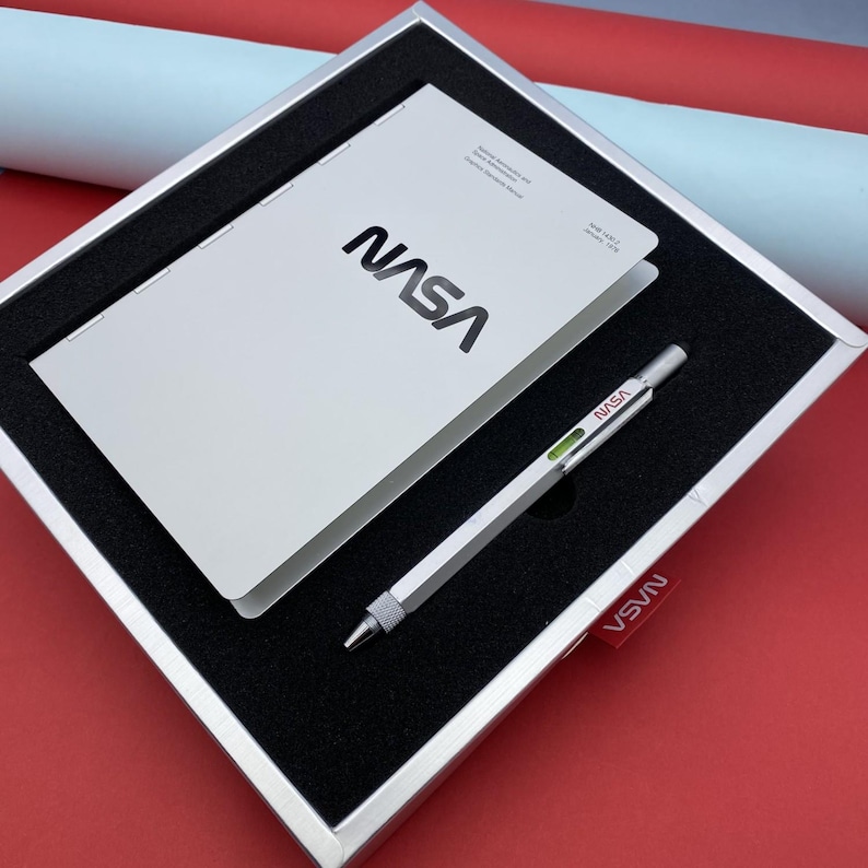 May include: A white gift box with a black interior. The box contains a white notebook with the NASA logo and a silver pen with a green level.