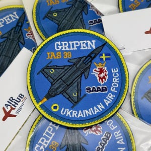 Puede incluir: Un parche bordado circular que presenta un avión de combate JAS 39 Gripen gris oscuro sobre un fondo azul. El texto dice GRIPEN JAS 39 y UKRAINIAN AIR FORCE. Incluye una bandera sueca, un emblema de león rojo y blanco y el logotipo SAAB en blanco.