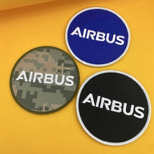 May include: Three circular patches featuring the word "AIRBUS" in white. One patch is blue, one is black, and the third has a green and brown camouflage design. Each patch has a white border.