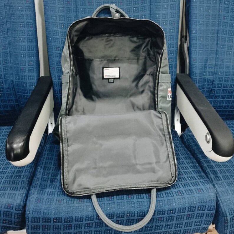 Puede incluir: Una mochila gris con un bolsillo frontal con cremallera y una cremallera negra. La mochila est&aacute; abierta y el interior es visible. La mochila est&aacute; sentada en un asiento con estampado azul y blanco.