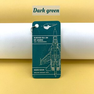 May include: A dark green keychain featuring a detailed outline of a Sukhoi SU-34 fighter jet with the text "SUKHOI SU-34", "RF-81852", "MSN: 4160662810801", "0220/1034", and "GENUINE AIRCRAFT SKIN".