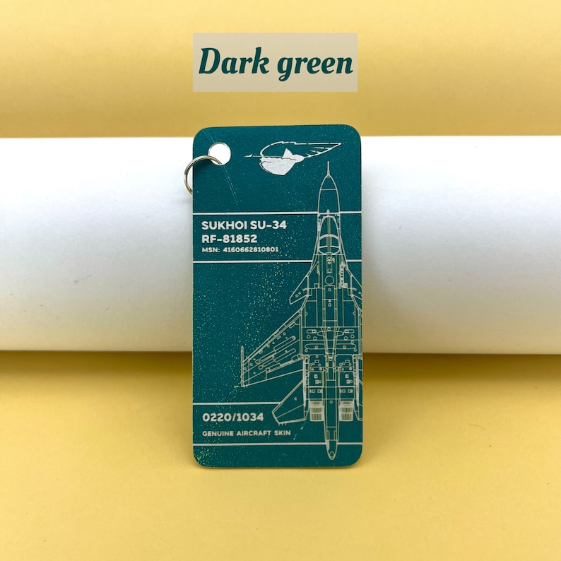 May include: A dark green keychain featuring a detailed outline of a Sukhoi SU-34 fighter jet with the text "SUKHOI SU-34", "RF-81852", "MSN: 4160662810801", "0220/1034", and "GENUINE AIRCRAFT SKIN".
