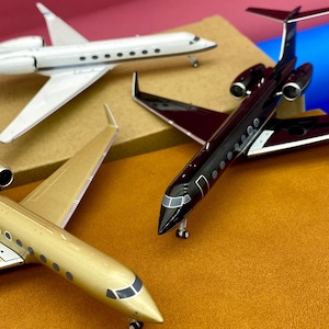 Könnte beinhalten: Vier Miniatur-Modellflugzeuge in Gold, Schwarz und Weiß. Das goldene Flugzeug befindet sich im Vordergrund, dahinter ein weißes und ein schwarzes Flugzeug. Die Flugzeuge haben detaillierte Merkmale, einschließlich Fenster und Flügel.