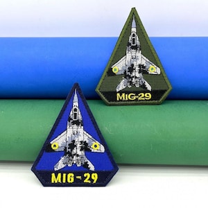 Può includere: Due toppe ricamate a forma di triangolo, ciascuna con un aereo da combattimento MiG-29 bianco su uno sfondo blu e verde. Il testo "MIG-29" è ricamato in giallo su ogni toppa.