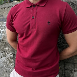 Könnte beinhalten: Ein rotes, kurzärmeliges Poloshirt mit einem kleinen Flugzeug-Design, das auf die Brust gestickt ist.