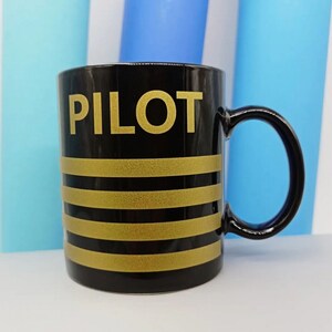 May include: Black ceramic mug with gold stripes and the word "PILOT" in gold lettering.