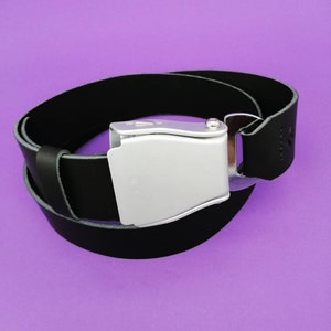 Könnte beinhalten: Ein schwarzer Ledergürtel mit einer silbernen Schnalle. Die Schnalle ist ein schlichtes, rechteckiges Design mit einer glatten, polierten Oberfläche.