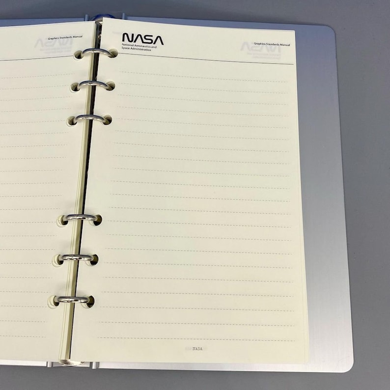 May include: A white notebook with a black and white NASA logo and the text "Graphics Standards Manual" on the cover. The notebook has a ring binder and is open to a blank page with lined paper.
